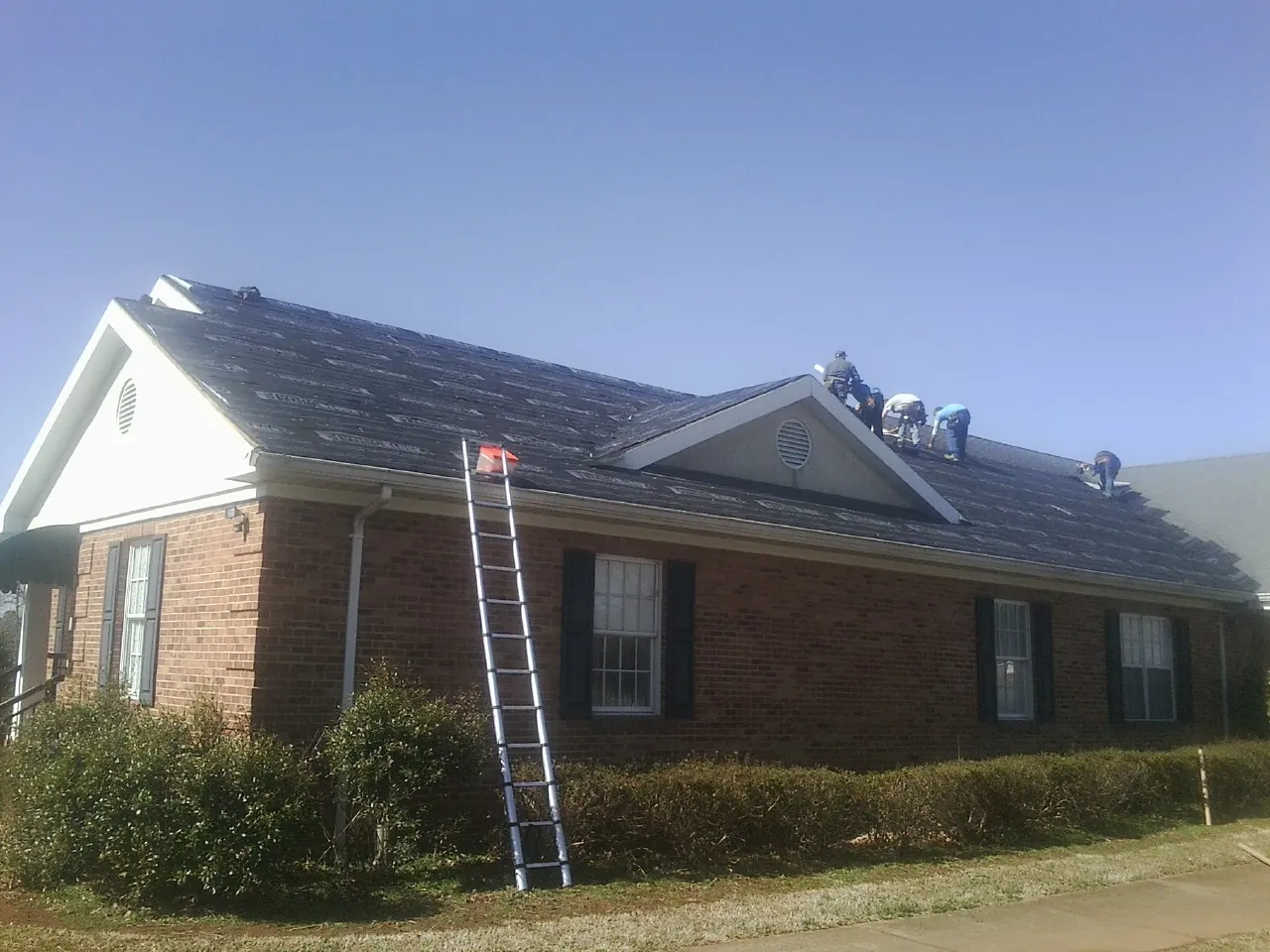Crew tearing shingles off a brick ranch roof with a ladder set — Metal Roof Repair work by our crew in Wilbraham