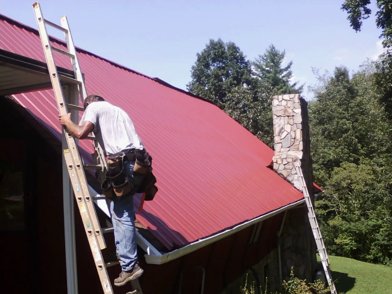 Roofer on a ladder beside a red metal roof and stone chimney — Soffit & Fascia Repair by Roofing Brothers in Wilbraham